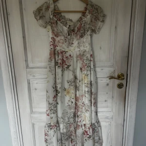 Blommig offshoulder långklänning - Romantisk långklänning med blommigt mönster i pastelliga toner. Offshoulder-modell med volangdetaljer och knytning framtill. Klänningen har en luftig passform och är perfekt för sommarens tillfällen. Helt ny med prislapp kvar 🌸🌸☀️