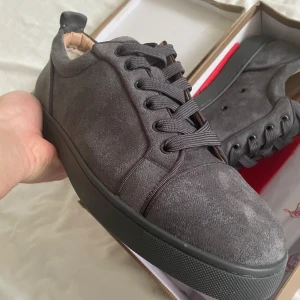 Louboutin  - Säljer ett par gråa sneakers från Christian Louboutin med klassisk röd sula och ovandel i mocka. Skorna har rund tå, svarta snören och diskret logga på insidan. Perfekta för dig som vill ha något stilrent men ändå lyxigt. 