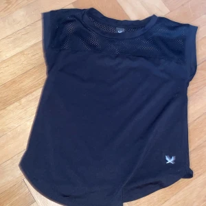 Svart sportig t-shirt från SOC - Svart t-shirt från SOC med meshdetaljer upptill för extra ventilation. Perfekt för träning eller aktiva dagar. Klassisk rund hals och ärmlös design ger en sportig look.