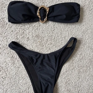 Svart bandeau bikini med gulddetalj - Säljer en svart bandeau bikini med snygg guldfärgad metallring framtill. Överdelen är axelbandslös och har knytning i ryggen. Oanvänd och i storlek S. 