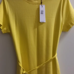 Gul t-shirtklänning från Calvin Klein - Säljer en gul t-shirtklänning från Calvin Klein med rund halsringning och korta ärmar. Klänningen har ett smalt bälte i midjan och diskret logga framtill. Perfekt för en avslappnad och trendig look.