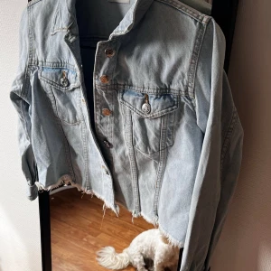 Ljusblå croppad jeansjacka - Säljer en ljusblå croppad jeansjacka med rå kant nertill och klassiska bröstfickor med knappar. Jackan har silverfärgade knappar framtill och vid ärmslut. Perfekt för dig som gillar en avslappnad och trendig stil.