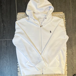 Ralph Lauren Zip Hoodie Vit  - Vit Ralph Lauren zip hoodie, storlek M, i fint skick trots att den är använd. Klassisk och stilren modell med dragkedja och det ikoniska lilla broderade logot på bröstet. Snörena i huvan saknas, men det påverkar inte funktionen eller looken nämnvärt. Skön och tidlös hoodie – perfekt för vardag och avslappnade tillfällen. 
