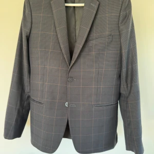 Grå/blå rutig kostym med kavaj och byxor - Säljer en stilren skräddarsydd blågrå kostym i rutigt mönster med matchande kavaj och byxor. Kavajen har smal passform, två knappar framtill, fyra knappar vid ärmslut och klassiska fickor. Perfekt för bal eller fest. Passar man som är drygt 180 till drygt 190 cm lång och har smal kroppsbyggnad. Storleken är uppskattad då kostymen är skräddarsydd. Byxorna är i 100-längd, så runt 150.