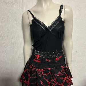 Svart och röd punkkjol från Myd Punks - Cool svart minikjol med röda kedje- och texttryck, säkerhetsnålsdetaljer och spetskant. Kjolen har ett punkigt uttryck med plisserad form och dekorativa detaljer framtill. Perfekt för dig som gillar alternativ stil.