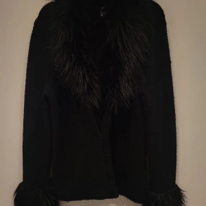 Svart kofta med fuskpäls från H&M - Säljer en svart stickad kofta från H&M med fuskpälsdetaljer vid krage och ärmslut. Koftan har långa ärmar och stängs med tryckknappar framtill. Perfekt för dig som vill ha en mysig och snygg look.