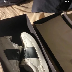 Valentino sneakers med svart detalj - Säljer ett par vita Valentino sneakers med bred svart rem över mitten. Skorna har klassisk rund tå, vita snören och diskret logga på plösen. Perfekta för dig som gillar stilrena och lyxiga sneakers med en twist. Dustbag och låda kommer med!