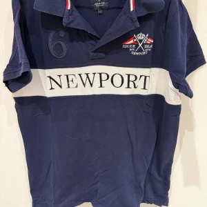 Marinblå pikétröja från Newport - Säljer en marinblå pikétröja från Newport med vit rand och broderad logga på bröstet. Tröjan har klassisk krage med röd och vit detalj samt siffran 6 broderad på axeln. Perfekt för en sportig och stilren look.