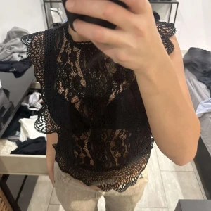 Svart spetsblus med volangdetaljer - Säljer en svart blus i spets med vackra volangdetaljer längs ärmar och nederkant. Blusen är ärmlös och har ett genombrutet mönster som ger en elegant look. Perfekt att styla till både jeans och kjol.