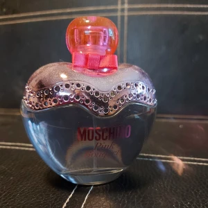 Moschino Pink Bouquet parfym - En hjärtformad parfymflaska från Moschino Pink Bouquet med glittrande dekorationer och rosa detaljer. Flaskan har en rosa kork och en dekorativ rosett vid halsen. Perfekt för dig som gillar söta och lekfulla dofter.