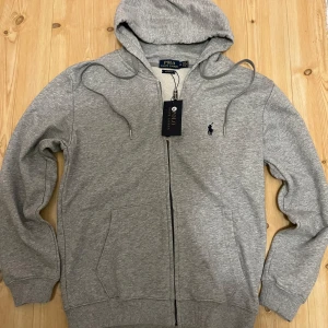 Grå hoodie från Polo Ralph Lauren - Säljer en klassisk grå hoodie från Polo Ralph Lauren i storlek M. Tröjan har dragkedja, huva med dragsko och två fickor framtill. Liten broderad logga på bröstet. Perfekt för en avslappnad stil.