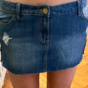 Blå jeanskjol med nitar i stl S - Säljer en blå jeanskjol med slitna detaljer och nitar på ena fickan. Kjolen har klassiska fickor fram och bak, bälteshällor och rå kant nertill. Perfekt för dig som gillar en edgy och trendig stil.