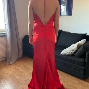 Röd långklänning med öppen rygg och slits - Säljer en röd långklänning med halterneck och djup öppen rygg. Klänningen har en smickrande drapering framtill och en hög slits på sidan. Perfekt för bal eller fest. Materialet är glansigt och följsamt. Snöret ska knytas där bak 