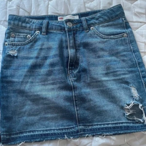Blå jeanskjol från Levi's - Säljer en klassisk blå jeanskjol från Levi's med slitna detaljer och rå kant nertill. Kjolen har fem fickor, bälteshällor och dragkedja framtill. Perfekt för en avslappnad och cool stil.