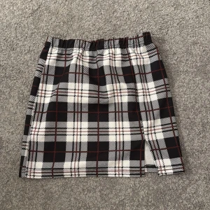 Rutig kortkjol med slits - Snygg kortkjol med svart, vit och röd rutig design. Kjolen har en liten slits framtill och resår i midjan för bekväm passform. Perfekt att styla med en enkel topp eller stickad tröja.