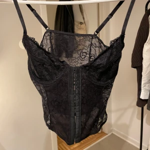 Svart spetskorsett med smala axelband - Säljer en svart korsett-topp i spets med smala justerbara axelband och dragkedja framtill. Toppen har en feminin och figurnära passform, perfekt att styla till olika outfits.