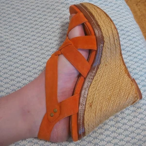 Orangea sandaler med kilklack - Säljer ett par snygga orangea sandaler med hög kilklack i rep och breda remmar i mocka. Perfekta för sommaren och ger en bohemisk känsla till outfiten. Lätta att ta på och av tack vare öppen design.