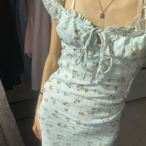 Ljusblå blommig klänning med volang och knyt - Söt ljusblå klänning med små blommor, volang vid bysten och knyt framtill. Klänningen har smala axelband och en figurnära passform. Perfekt för varma dagar och skolavslutningen. Har använt 1 gång.