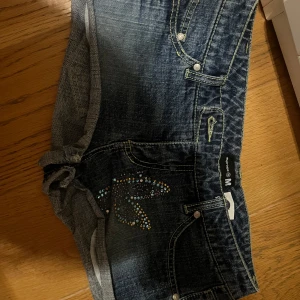 Jeansshorts med fjärilsdetalj i strass - Mörkblå jeansshorts  med en stor fjäril i färgglada strasstenar på baksidan och mindre strasstenar framtill. Klassisk femficksmodell med låg midja och korta ben. Perfekta för sommaren! Jag har storlek 36 och de passar perfekt på mig 