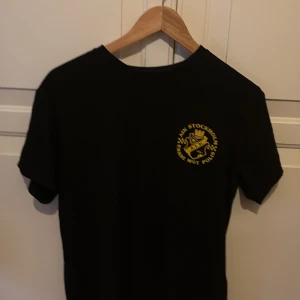 Svart AIK t-shirt med gul text - Svart t-shirt med gul AIK-logga och texten 'AIK Skapade Mot Polis' tryckt på bröstet och ryggen. Klassisk passform och rund halsringning. Perfekt för dig som vill visa ditt AIK-stöd.