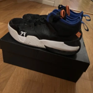 Svarta Jordan sneakers med blå och orange detaljer - Säljer ett par svarta Jordan sneakers med vita sulor, blå insida och orange detaljer. Skorna har nummer 23 på sidan och Jumpman-loggan under sulan. Perfekta för dig som gillar basketstil och vill sticka ut med färgglada detaljer.