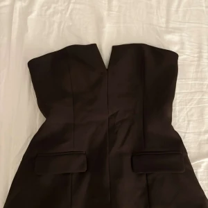 Svart axelbandslös klänning - Säljer en svart axelbandslös klänning med figurnära passform och markerad midja. Klänningen har två dekorativa ficklock framtill och en stilren, minimalistisk design. Perfekt för dig som gillar en klassisk och elegant look.