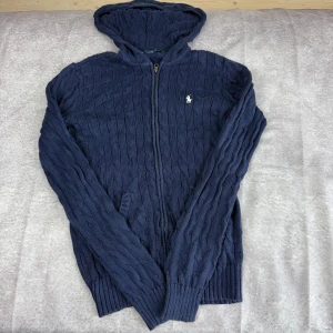 Marinblå stickad hoodie från Ralph Lauren - Säljer en marinblå stickad hoodie från Ralph Lauren med dragkedja framtill och kabelstickat mönster. Tröjan har huva, långa ärmar och en broderad logga på bröstet. Perfekt för kyliga dagar.