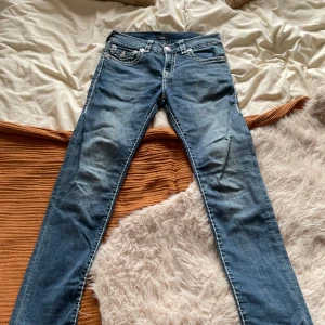 Blå jeans från True Religion med vita sömmar - Säljer ett par blå jeans från True Religion med tydliga vita kontrastsömmar och snygga fickdetaljer bak. Klassisk femficksmodell med raka ben och låg midja. Märkespatch bak i linningen.