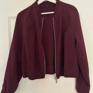 Vinröd bomberjacka med dragkedja - Säljer en vinröd bomberjacka med silverfärgad dragkedja framtill. Jackan har lång ärm, ribbade muddar och en klassisk bombermodell. Perfekt för dig som vill ha en stilren och enkel jacka till vardags.
