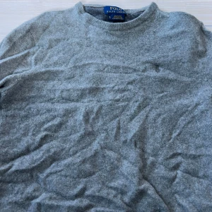 Grå ulltröja från Polo Ralph Lauren - Mysig grå tröja från Polo Ralph Lauren i mjuk ull. Klassisk rund halsringning och lång ärm, perfekt för kyliga dagar. Enkel och stilren design som passar till det mesta. Storleken är M
