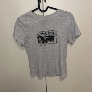 Grå t-shirt med New York-tryck - Säljer en ljusgrå t-shirt med ett svartvitt tryck av Downtown New York och en pizzeria på bröstet. T-shirten har rund hals och korta ärmar, perfekt för en sommaren 🫶🏻