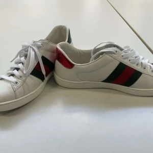 Gucci Ace sneakers med bi-detalj - Säljer ett par vita Gucci Ace sneakers med klassiska gröna och röda ränder på sidorna samt broderad guldfärgad bi. Baksidan har en röd och en grön metallic detalj med Gucci-logga. Skorna har rund tå och snörning. Priset är ej hugget i sten |ÄKTA✅|