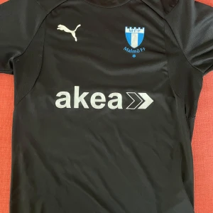 Malmö FF fotbollströja - Säljer en svart Malmö FF fotbollströja från Puma med klubbmärke och sponsortryck på bröstet. Tröjan har korta ärmar och är tillverkad i ett lätt och sportigt material, perfekt för träning eller en trendig look.