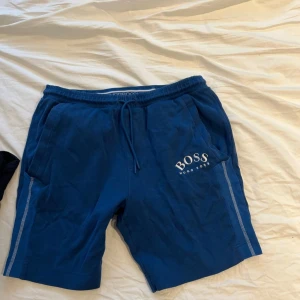 Blå shorts från Hugo Boss - Snygga blå shorts från Hugo Boss med vit logga på vänster ben. Shortsen har snörning i midjan, sidofickor och dekorativa ränder längs sidorna. Perfekta för en avslappnad stil.