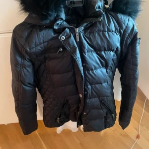 Svart jacka med pälshuva från Rockandblue - Snygg svart quiltad jacka från Rockandblue med pälskantad huva och flera dragkedjedetaljer. Jackan har lång ärm, markerad midja och praktiska fickor. Perfekt för kyligare dagar och ger en cool look.