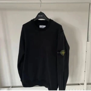 Svart tröja från Stone Island - Säljer en svart tröja från Stone Island med klassisk rund hals och den ikoniska loggan på ärmen. Tröjan är enkel och stilren, perfekt för dig som gillar streetwear och vill ha något bekvämt och snyggt.