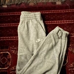 Grå mjukisbyxor från Nike - Säljer ett par klassiska grå mjukisbyxor från Nike med resår i midjan och fickor på sidorna. Byxorna har en enkel och sportig look med Nike-logga tryckt på låret. Perfekta för chill eller träning.