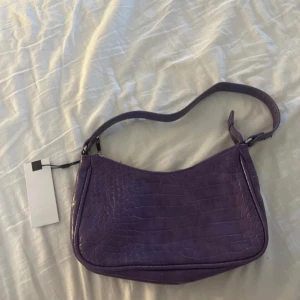  Axelväska  - Säljer en lila axelväska från  med krokopräglat mönster. Väskan har dragkedja i silver  upptill och ett stilrent, modernt utseende. Perfekt för att lyfta din outfit med en färgklick. Prislapp  sitter kvar.