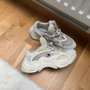 Chunky vita sneakers från Tommy Hilfiger - Säljer ett par chunky vita sneakers från Tommy Hilfiger med beige och grå detaljer. Skorna har snörning och den klassiska Tommy-loggan på sidan. Perfekta för en sportig och trendig look. 