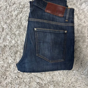 Mörkblå jeansbyxor från tiger of sweden - Snygga mörkblå jeans från Levis med klassisk femficksdesign och bruna detaljer på bakfickan. Jeansen har raka ben och tydliga kontrastsömmar. Perfekta för en stilren vardagslook.