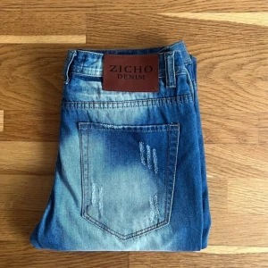  Zicho Denim-blåjeans-destroyed straight strl 33/32 - Ett par blå jeans från Zicho Denim med en unik och trendig design. Destroyed regular straight jeans. Perfekt kombination av stil och komfort. - 100% Bomull - Förslutning : knappgylf - Strl. : W33 L32