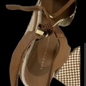 Bruna sandaler med knyt från Tommy Hilfiger - Snygga bruna sandaler från Tommy Hilfiger med knyt runt vristen och rutigt mönster på klacken. Skorna har öppen tå och detaljer i guld. Perfekta för en trendig look.