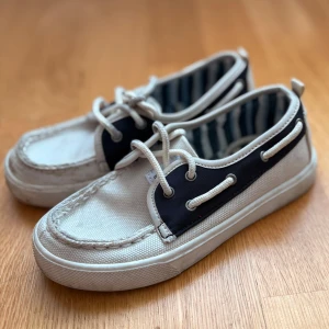 Vita och marinblå sneakers med snörning(barnskor) - Säljer ett par vita sneakers med marinblå detaljer och snörning. Skorna har en låg profil, rund tå och vit platt sula. Insidan är fodrad med randigt tyg i vitt och marinblått. Perfekta för en avslappnad stil. 