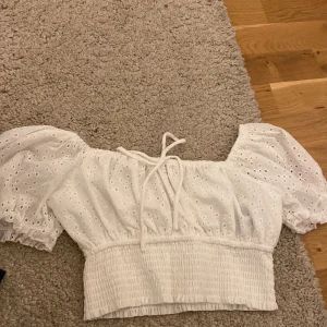 Vit broderad croppad blus från FB Sister - Superfin vit croppad blus från FB Sister med puffärmar och broderade hål. Toppen har knytning  och smockad midja för en snygg passform. Perfekt till sommaren!