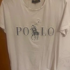 Vit t-shirt från Polo Ralph Lauren - Vit t-shirt från Polo Ralph Lauren med klassiskt POLO-tryck och broderad logga på bröstet. T-shirten har rund halsringning och korta ärmar. Perfekt för en stilren och sportig look.
