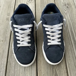 Mörkblå sneakers från Polo Ralph Lauren - Snygga mörkblå sneakers från Polo Ralph Lauren i strl: 40 med vit sula och vita skosnören. Baktill finns ett rutigt tyg i grönt, blått och gult samt Polo-logga. Skorna är i mocka och kommer med original snören. Nypris: 1499.