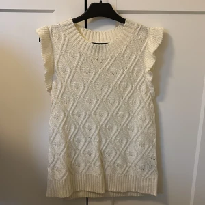 Vit stickad ärmlös topp med volang - Säljer en vit stickad topp med snyggt mönster och volangdetaljer vid axlarna. Toppen är ärmlös och har en rund halsringning samt ribbade kanter. Perfekt att styla över en skjorta eller t-shirt.