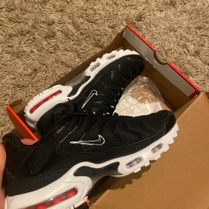 Nike Air Max Plus svarta sneakers - Lätt använda. Svarta Nike Air Max Plus sneakers med vit sula och röda detaljer i sulan. Skorna har tydliga luftbubblor, vit swoosh-logga på sidan och klassisk platt sula. Perfekta för dig som gillar sportig och modern stil.