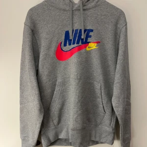 Grå Nike hoodie med färgglada loggor - Säljer en grå hoodie från Nike med stor färgglad logga framtill i blått, rött och gult. På ryggen finns tre mindre Nike-loggor i sammaj färger. Tröjan köptes in men användes aldrig.