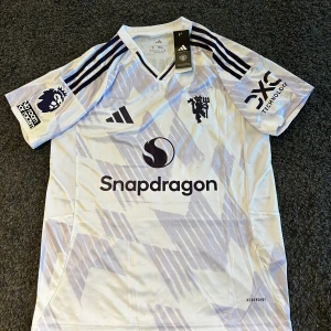 Manchester United 25/26 bortaställ - Manchester Uniteds bortatröja från Adidas med Cunha och nummer 10 på ryggen. Tröjan är vit med ljuslila grafiskt mönster, svarta detaljer och broderat klubbmärke. Tillverkad i lätt och ventilerande polyester med AEROREADY-teknologi. Klassisk V-ringning och korta ärmar. Helt ny en replika 